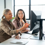 Smiling professionals in 30s and 50s, casually dressed, sitting at task table and discussing content on monitor.